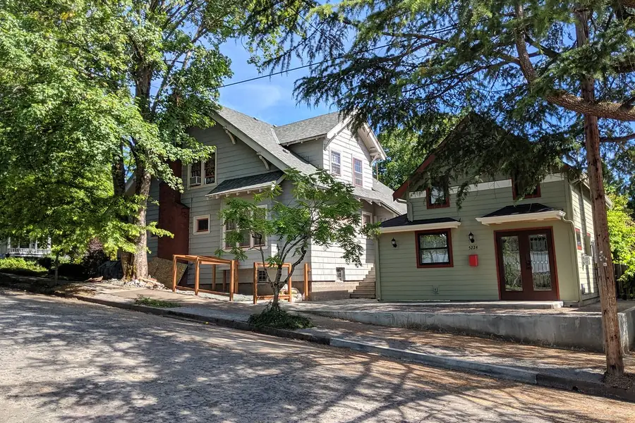 A small one-story cottage with green siding and a glass door sits beside a larger two-story family house under shade trees.