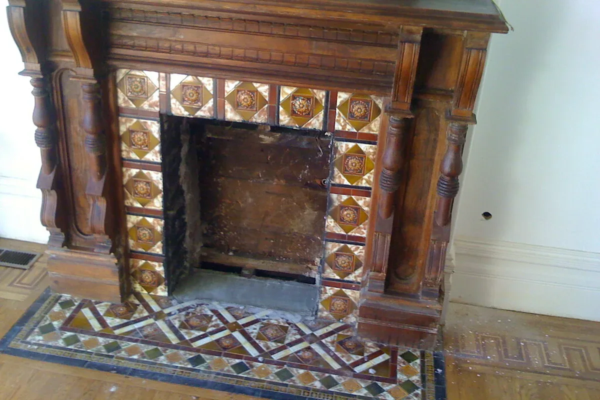 An old masonry fireplace with its brick firebox lining torn out, the bare opening framed by a carved wood mantel and a tiled hearth, ready for a gas insert.