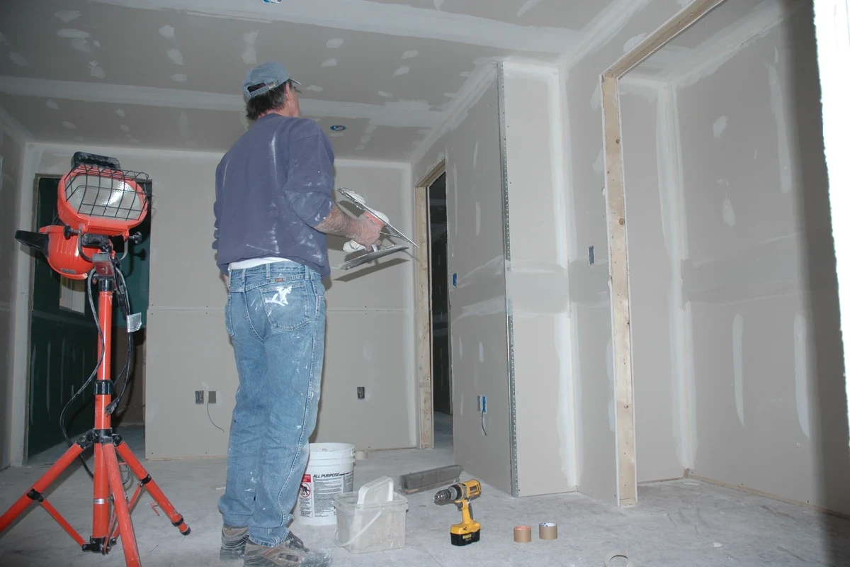 A worker in a cap and jeans tapes drywall seams in a stripped room, a work light and tools on the bare floor.